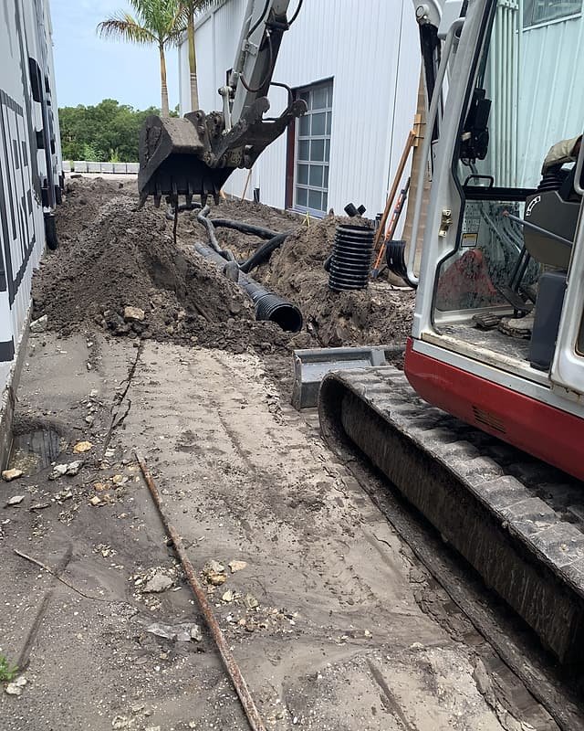 Compact excavator digging a drainage trench beside a building