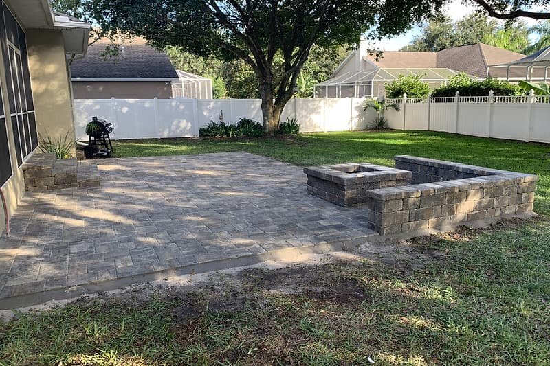 Paver patio extending into the lawn, with a fire pit and seating wall under shade trees