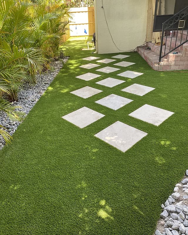 Side yard finished with artificial turf, square stepping stones and a white river-rock border