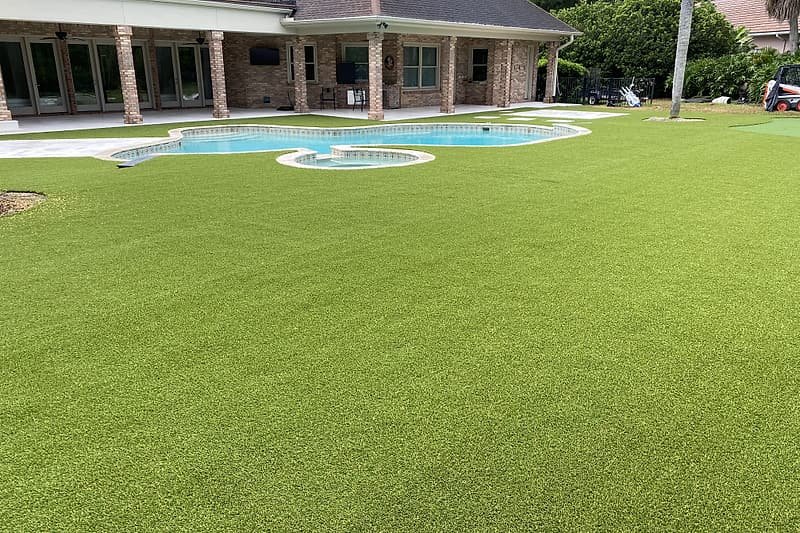 Large lawn of artificial turf stretching from the pool to the back of a two-story brick home
