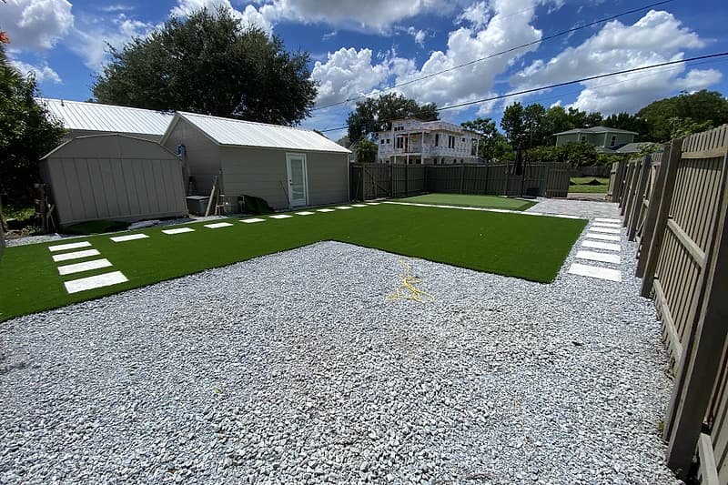 Backyard with an artificial turf lawn, gravel borders and a paver walkway along the fence